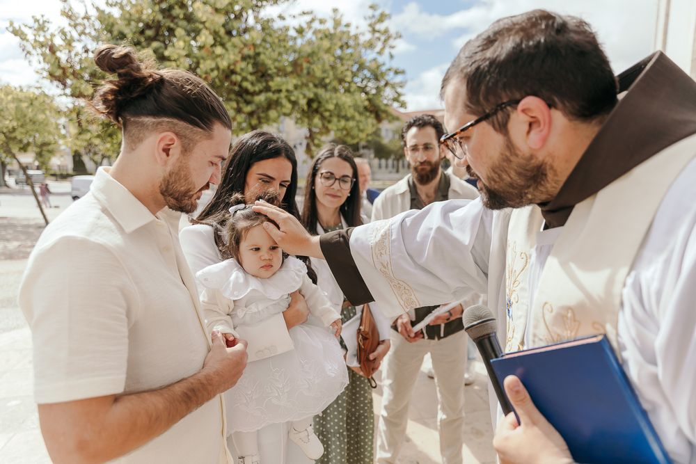 Priest performing a baptism ceremony on a baby held by parents outdoors on a sunny day.