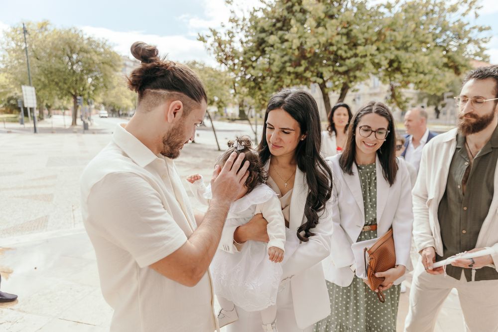 Group of people in white attire gathered outdoors, with a man holding a camera photographing a baby.