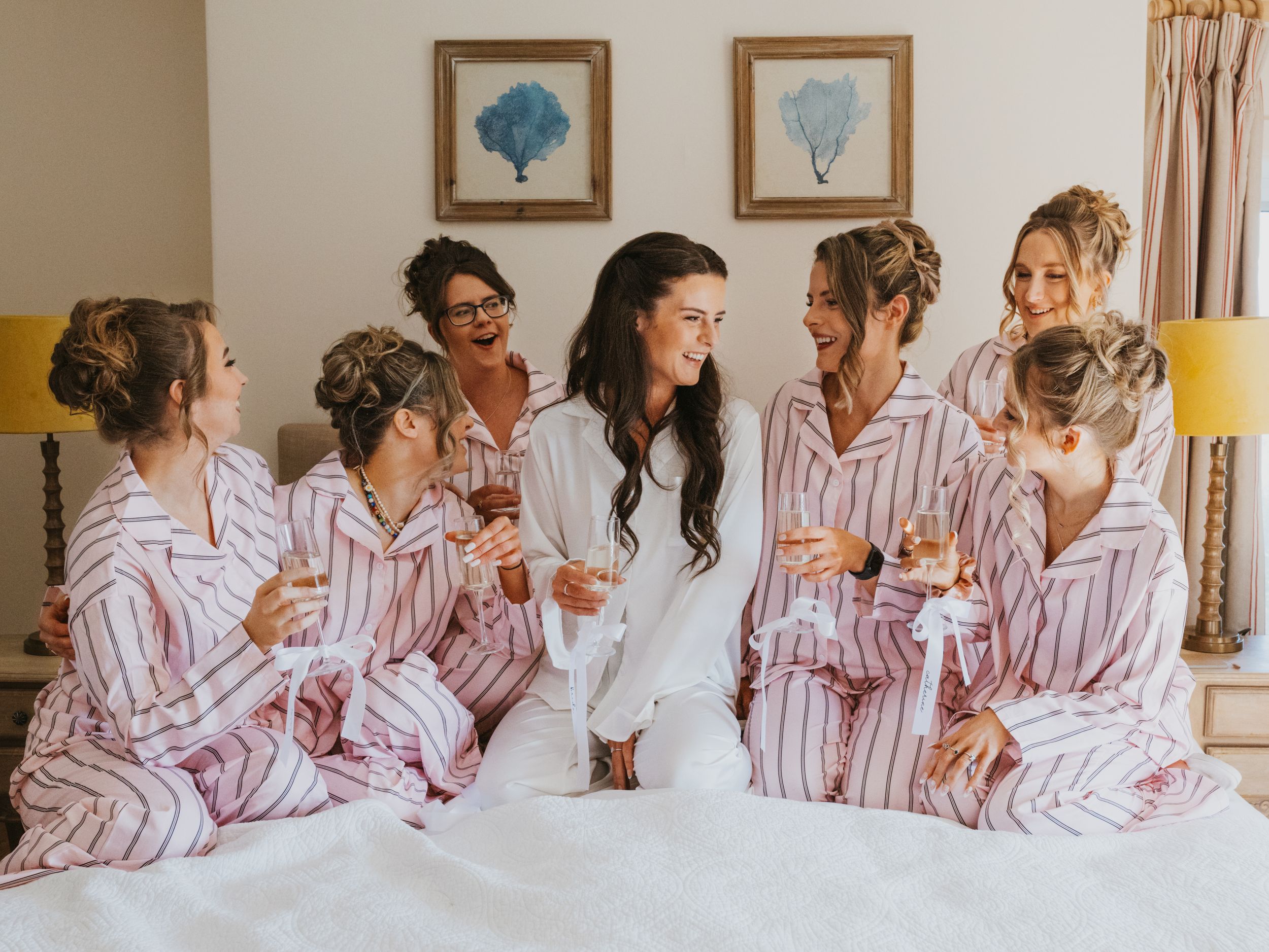 Bride and bridesmaids in matching pink striped pajamas sitting on bed laughing and holding champagne glasses.