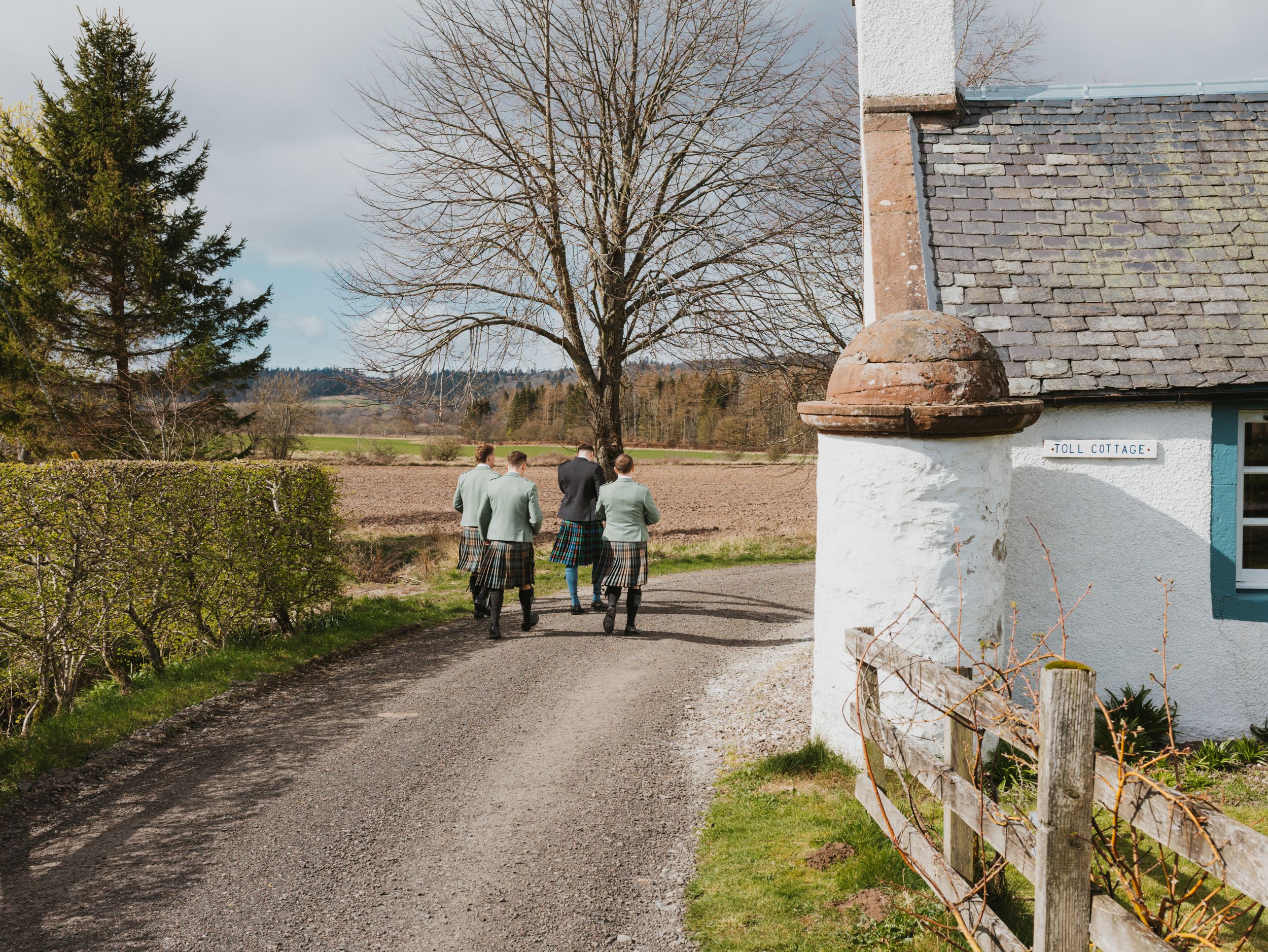 Group of people walking down a rural country lane past a white cottage with scenic farmland views.