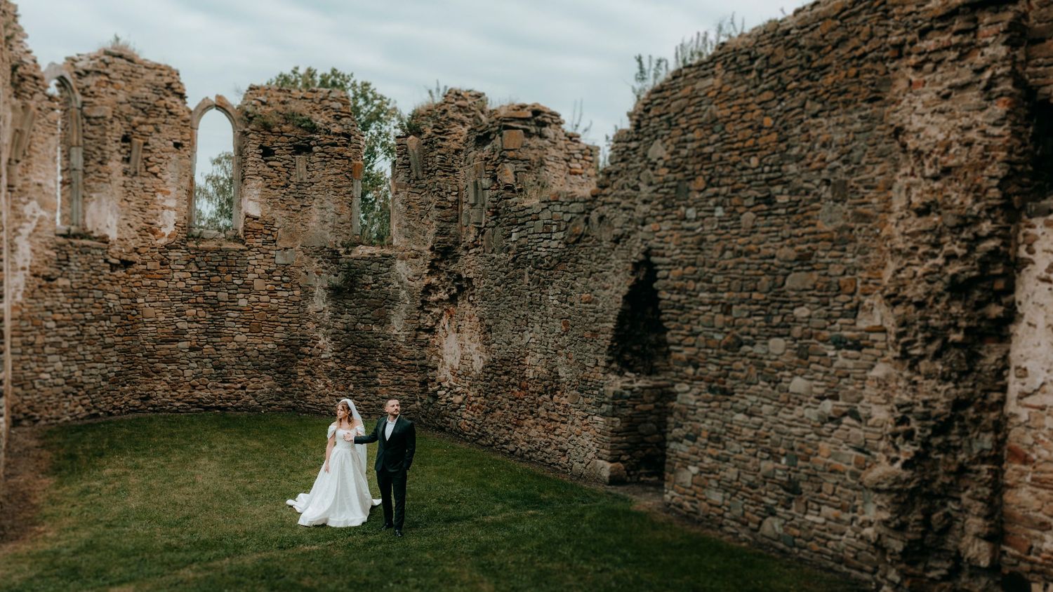 Bride and groom standing together inside ancient stone ruins surrounded by green grass.