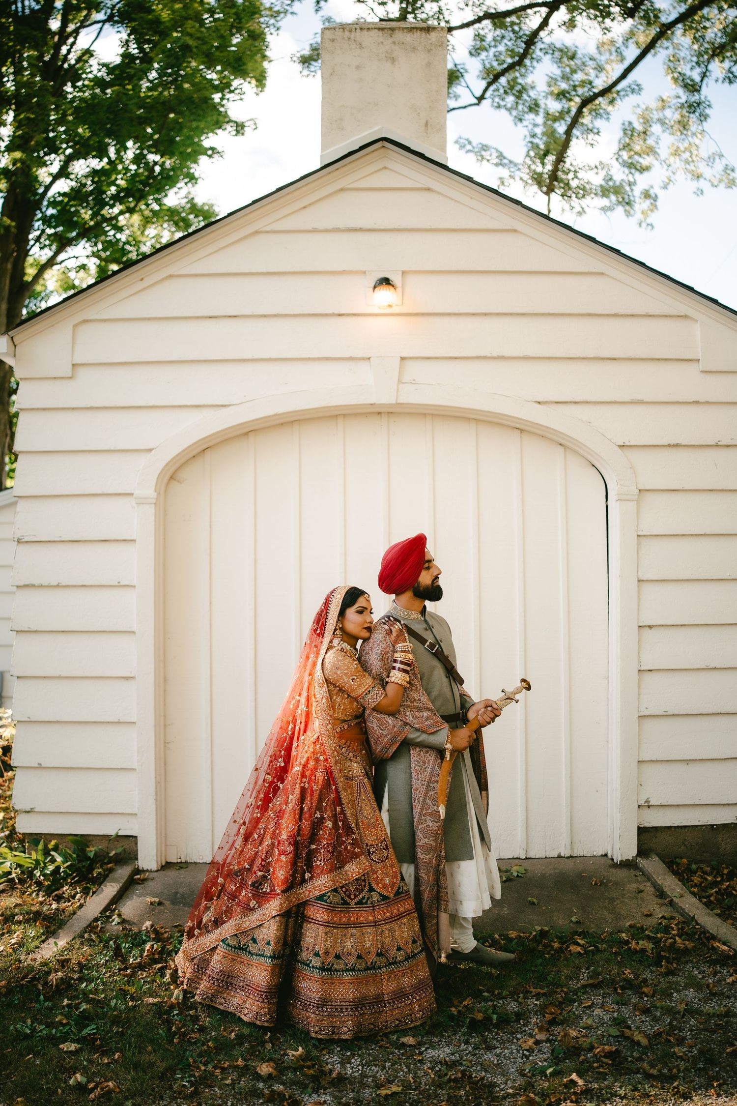 South Asian bride in red lehenga and groom in gray sherwani with red turban pose before white chapel doors.