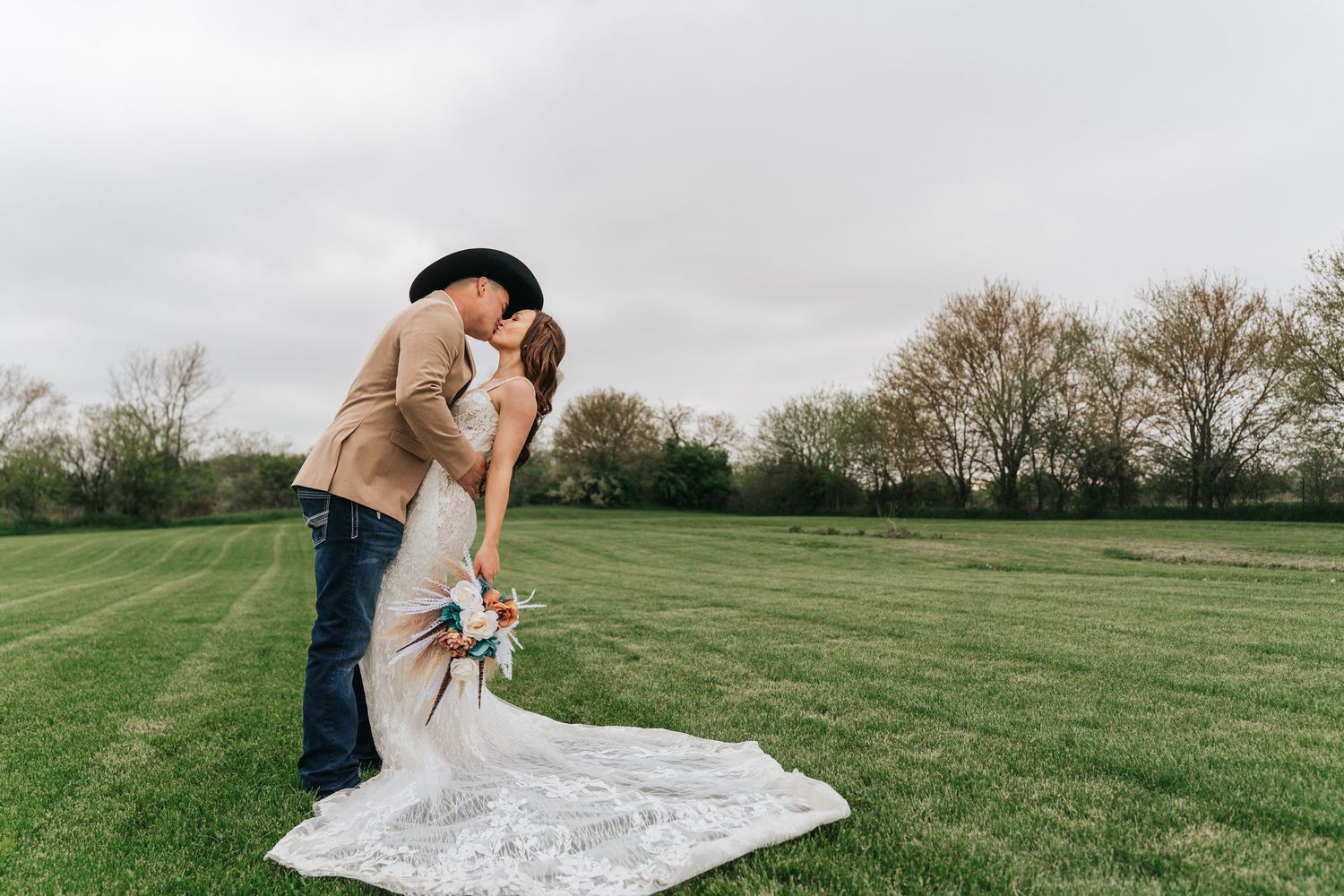 Couple sharing romantic dip kiss in green field, bride in lace gown with train, groom in cowboy hat.