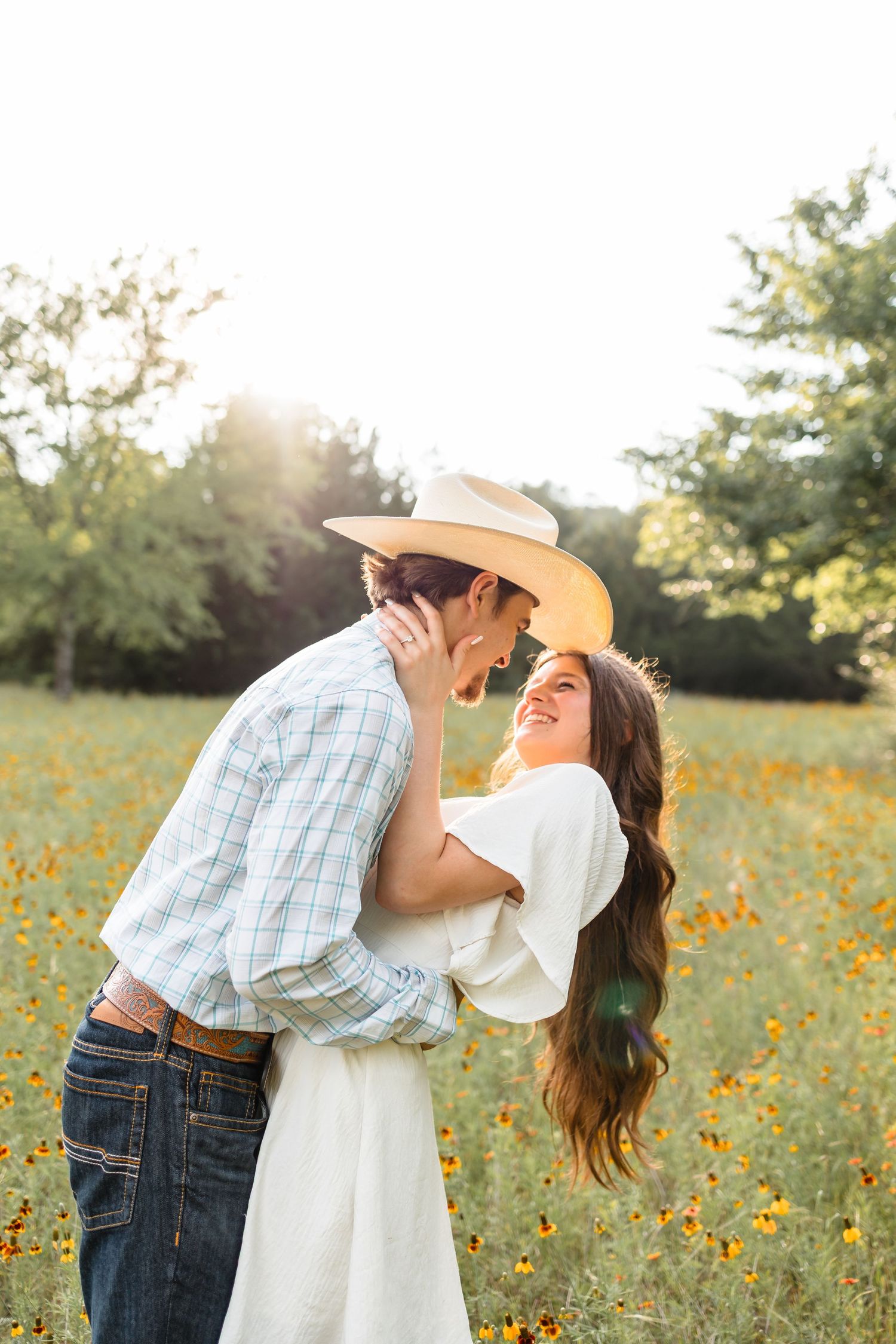 Couple sharing a romantic moment in a sunlit wildflower field, woman smiling in a cowboy hat.