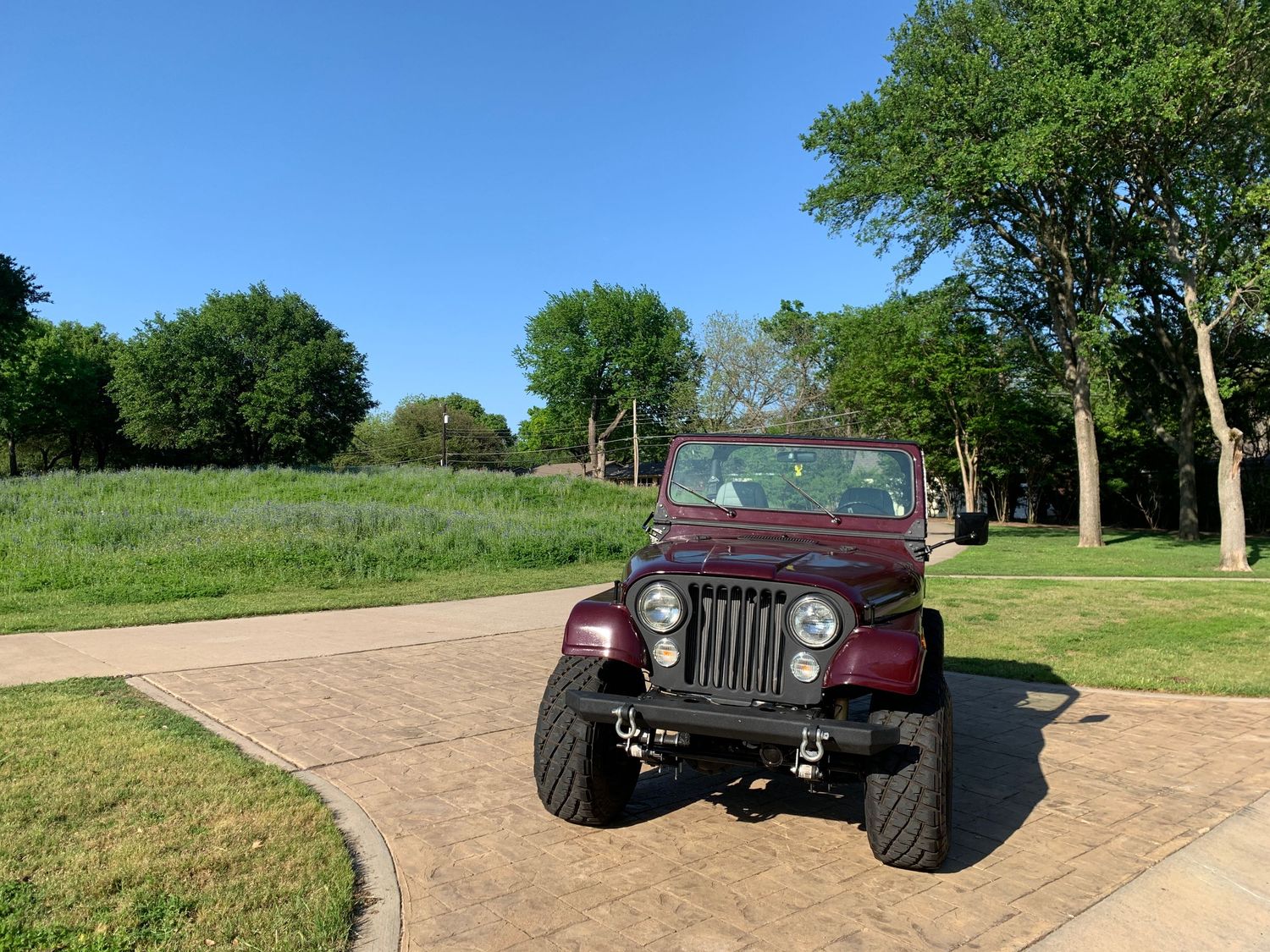Maroon 1986 Jeep CJ7 off-road vehicle parked on a driveway surrounded by green trees and blue wildflowers on a sunny day.