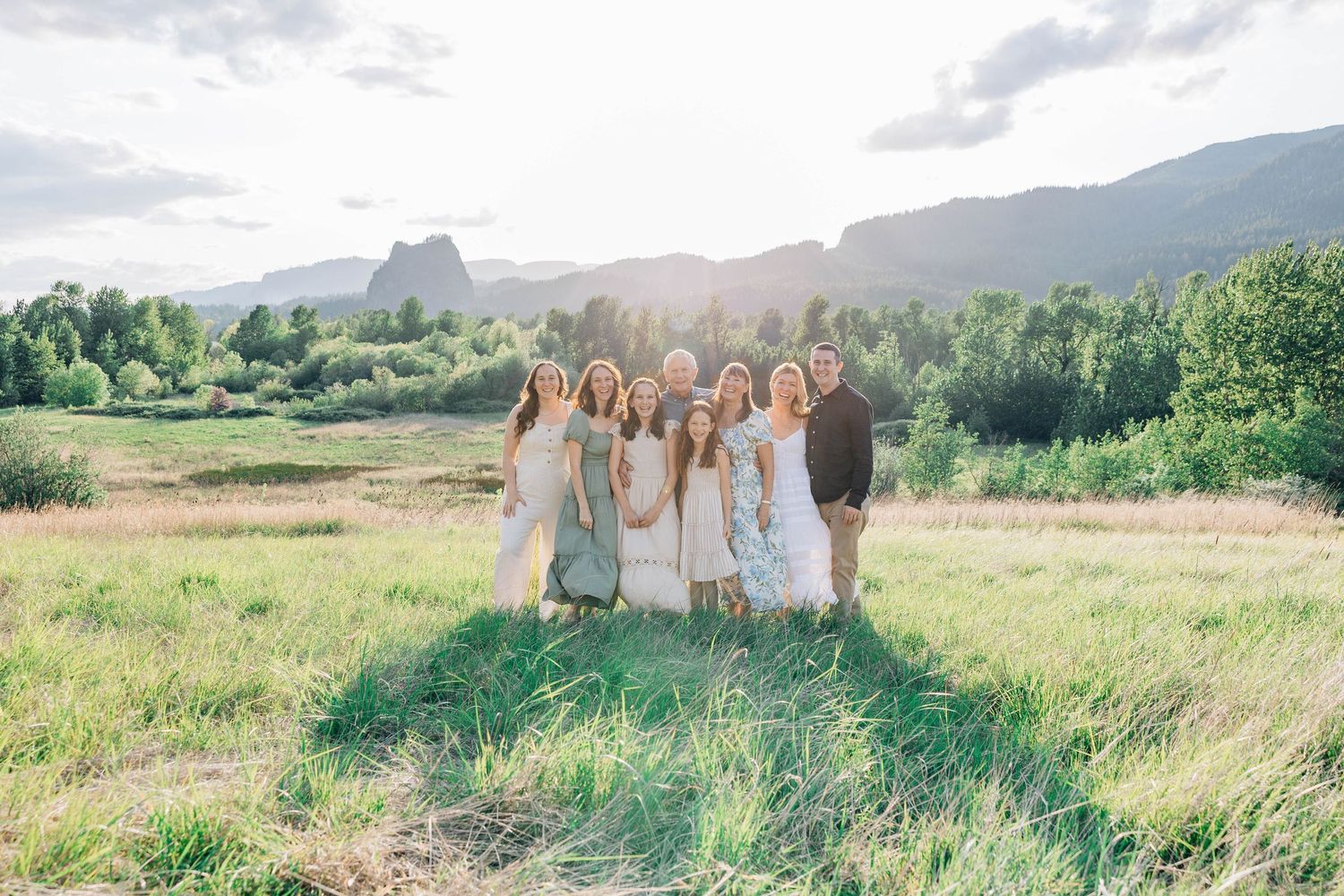 Family group photo in a green meadow with mountain backdrop during golden hour, wearing coordinated white and sage outfits.