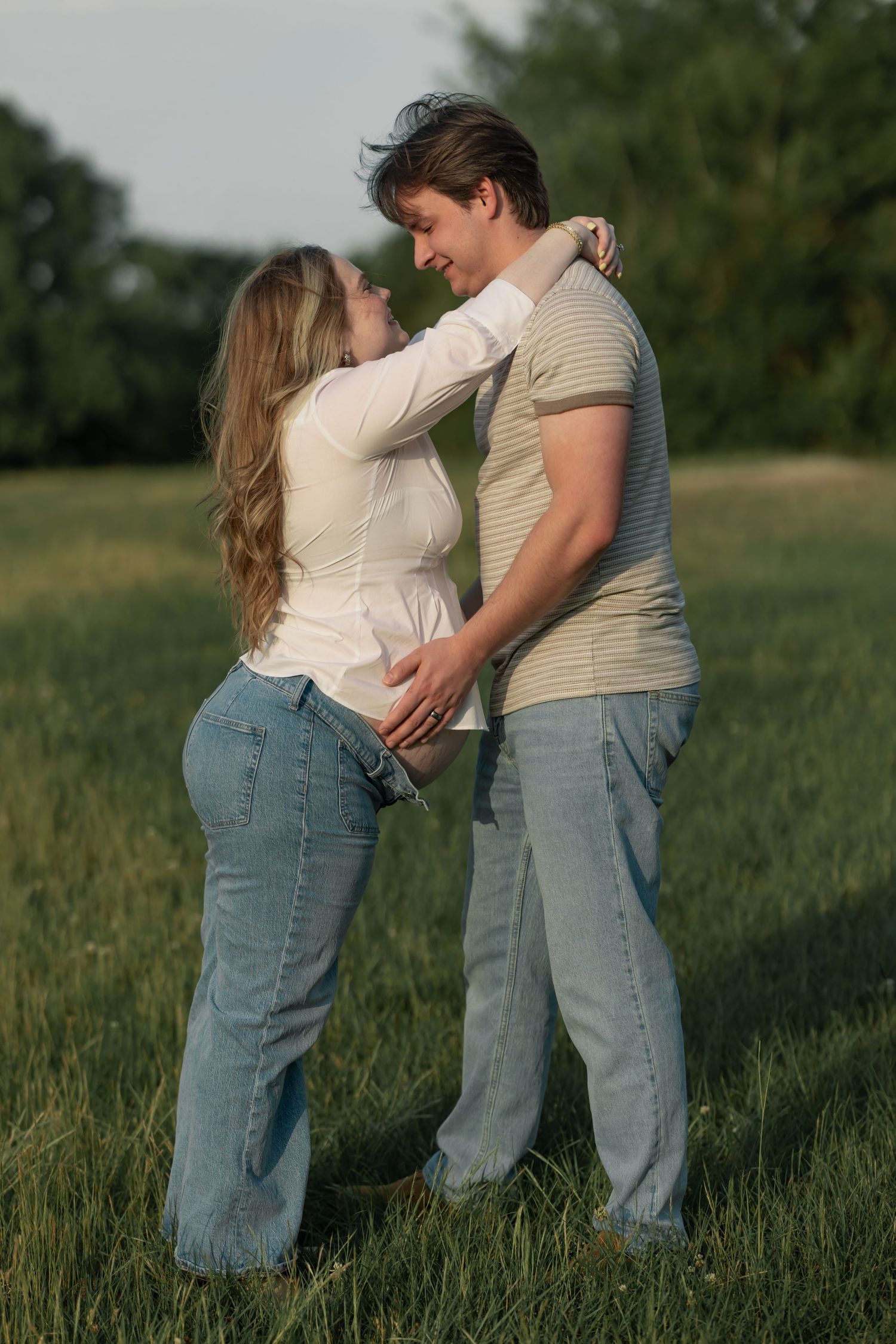 A couple embracing in a green field, smiling at each other, wearing jeans and casual tops on a sunny day.