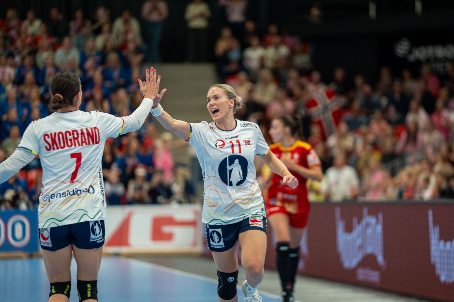 Two female handball players in white Norway jerseys celebrate with a high-five during a match.