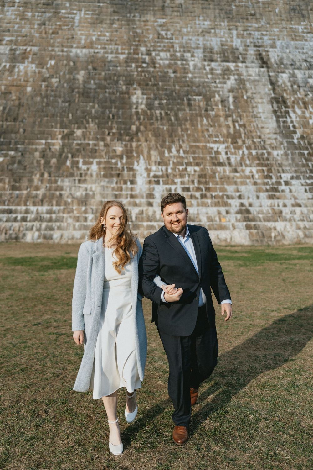 Smiling couple walking arm in arm outdoors near a large stone wall, woman in white dress and blue coat, man in dark suit.