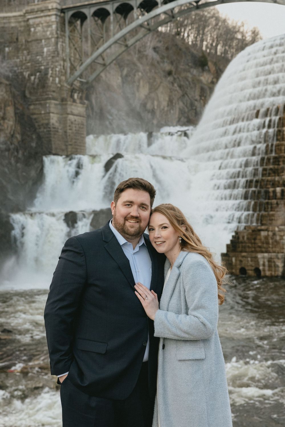 Couple smiling together in front of a stunning waterfall and stone arch bridge backdrop.