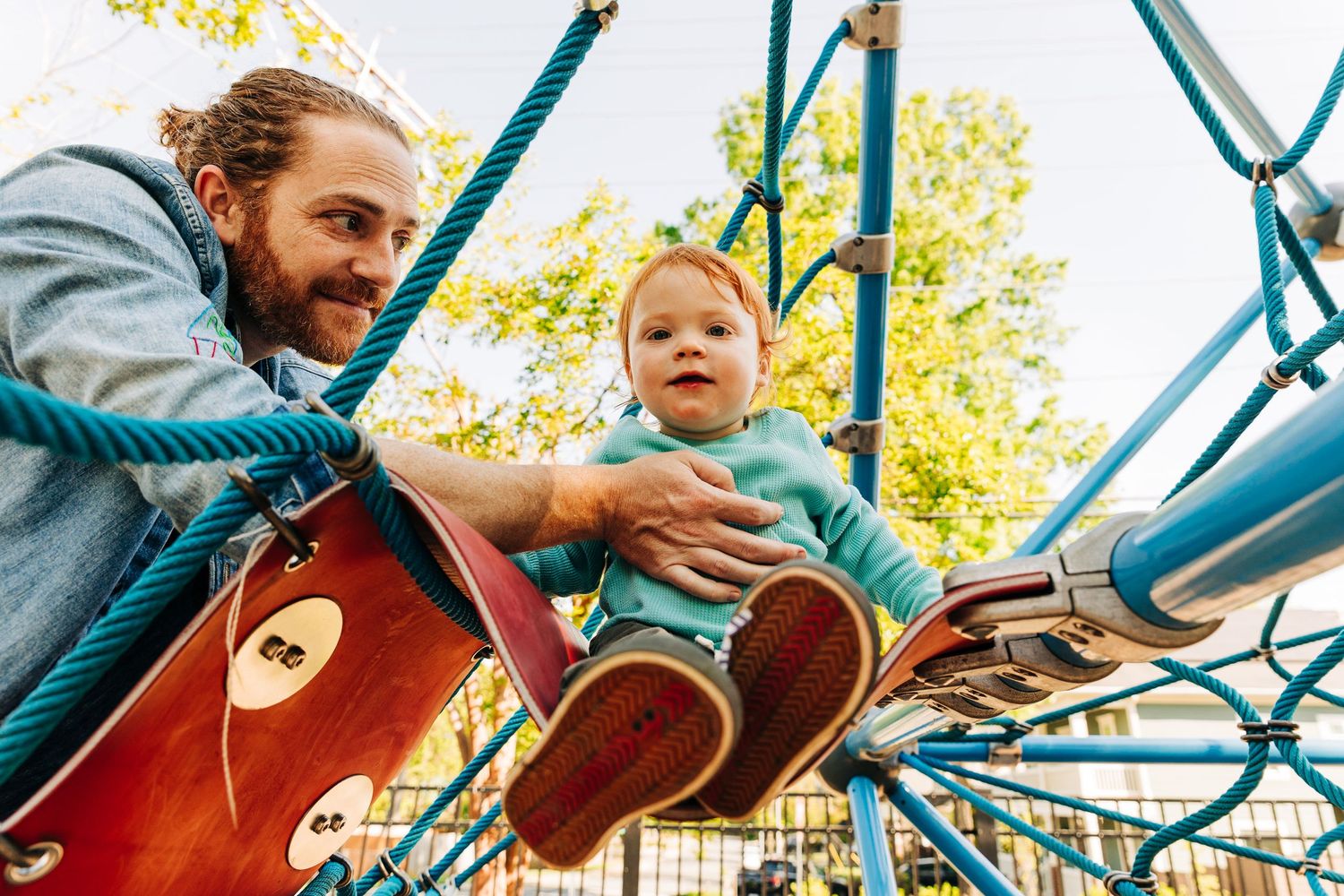 Father helping toddler play on colorful playground climbing structure outdoors on a sunny day.
