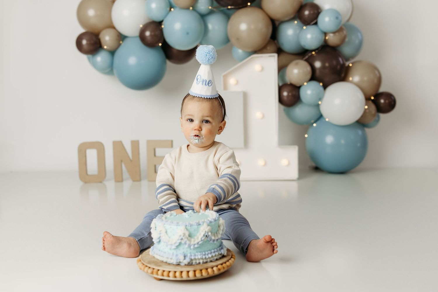 Baby in a party hat sits before a blue smash cake, surrounded by balloons and ONE sign at a first birthday celebration.