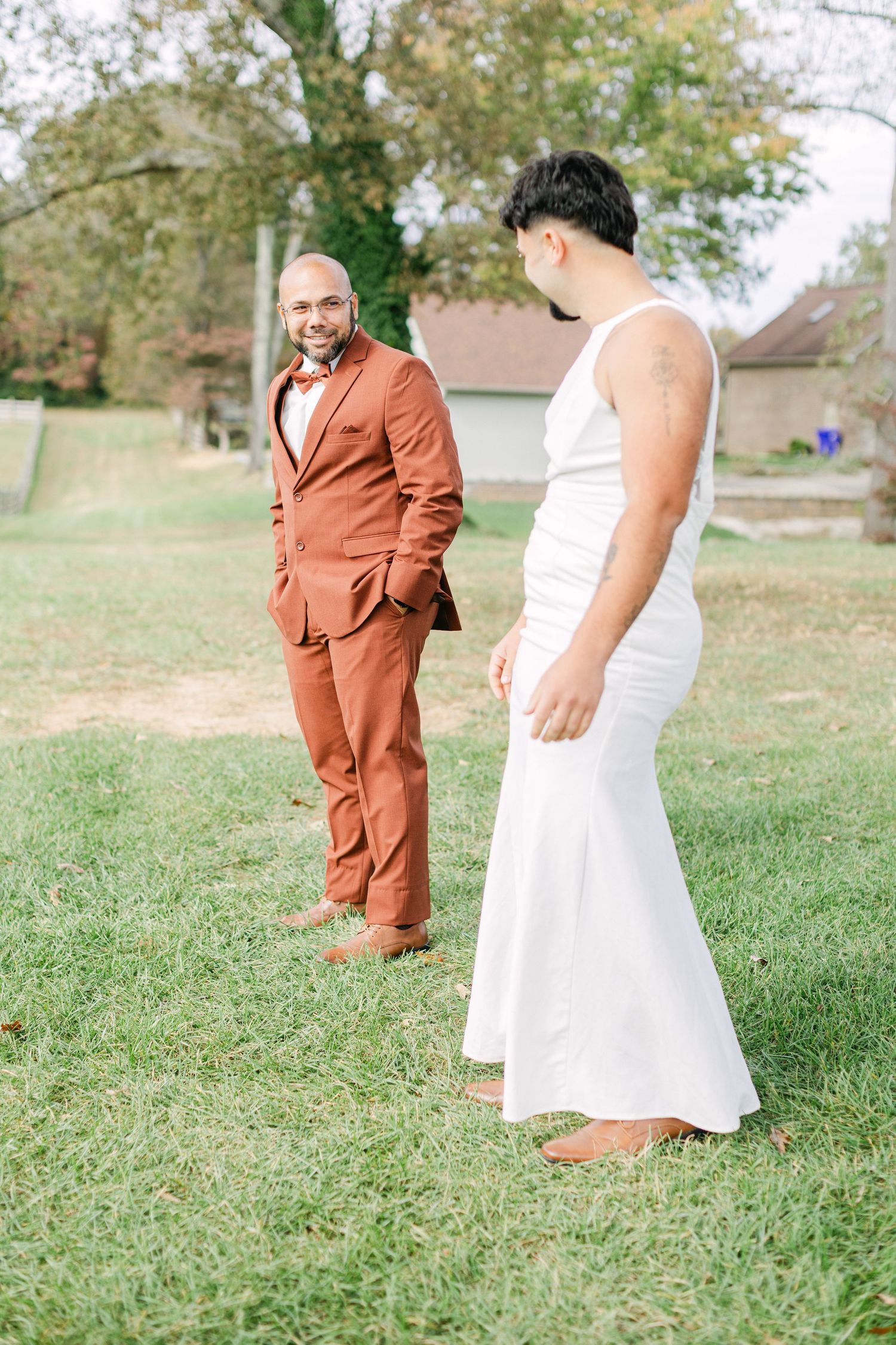 Two grooms share first look moment on green lawn, one in rust suit and one in white wedding dress.