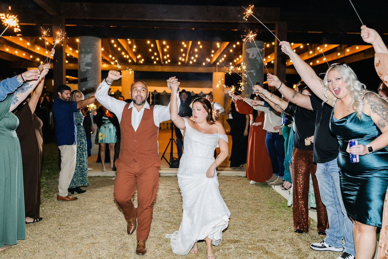 Bride and groom exit wedding reception through sparkler tunnel surrounded by cheering guests at night.