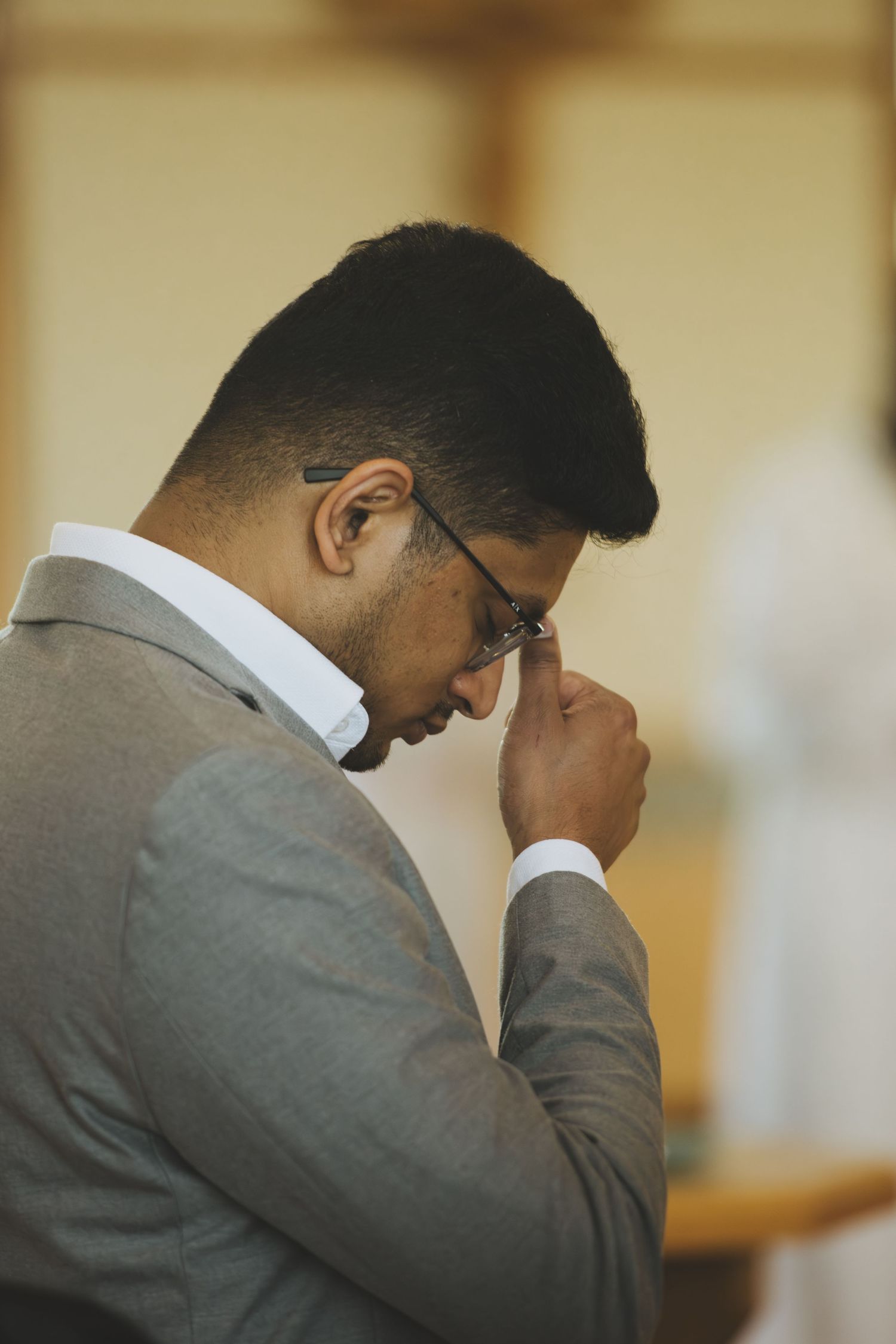 Man in gray suit bowing head in prayer inside a church, wearing glasses and adjusting them with one hand.