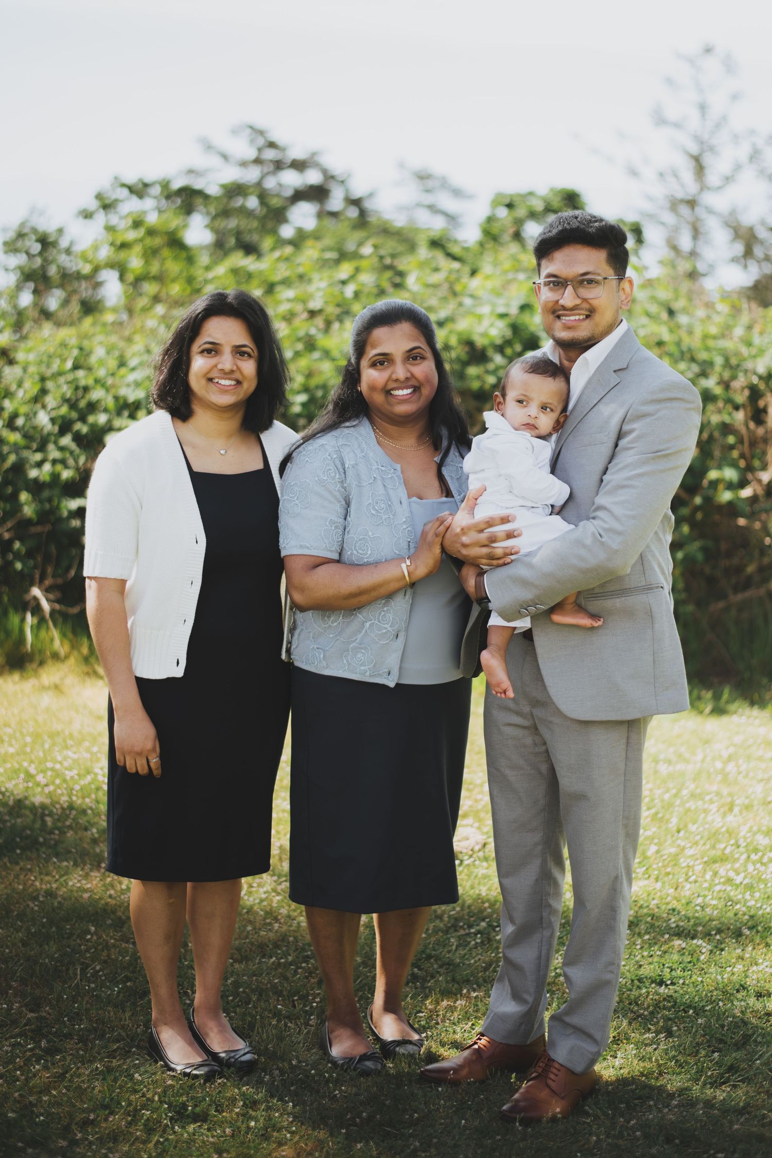 Family of four posing outdoors, man in gray suit holding baby, two women in formal attire, green trees background.