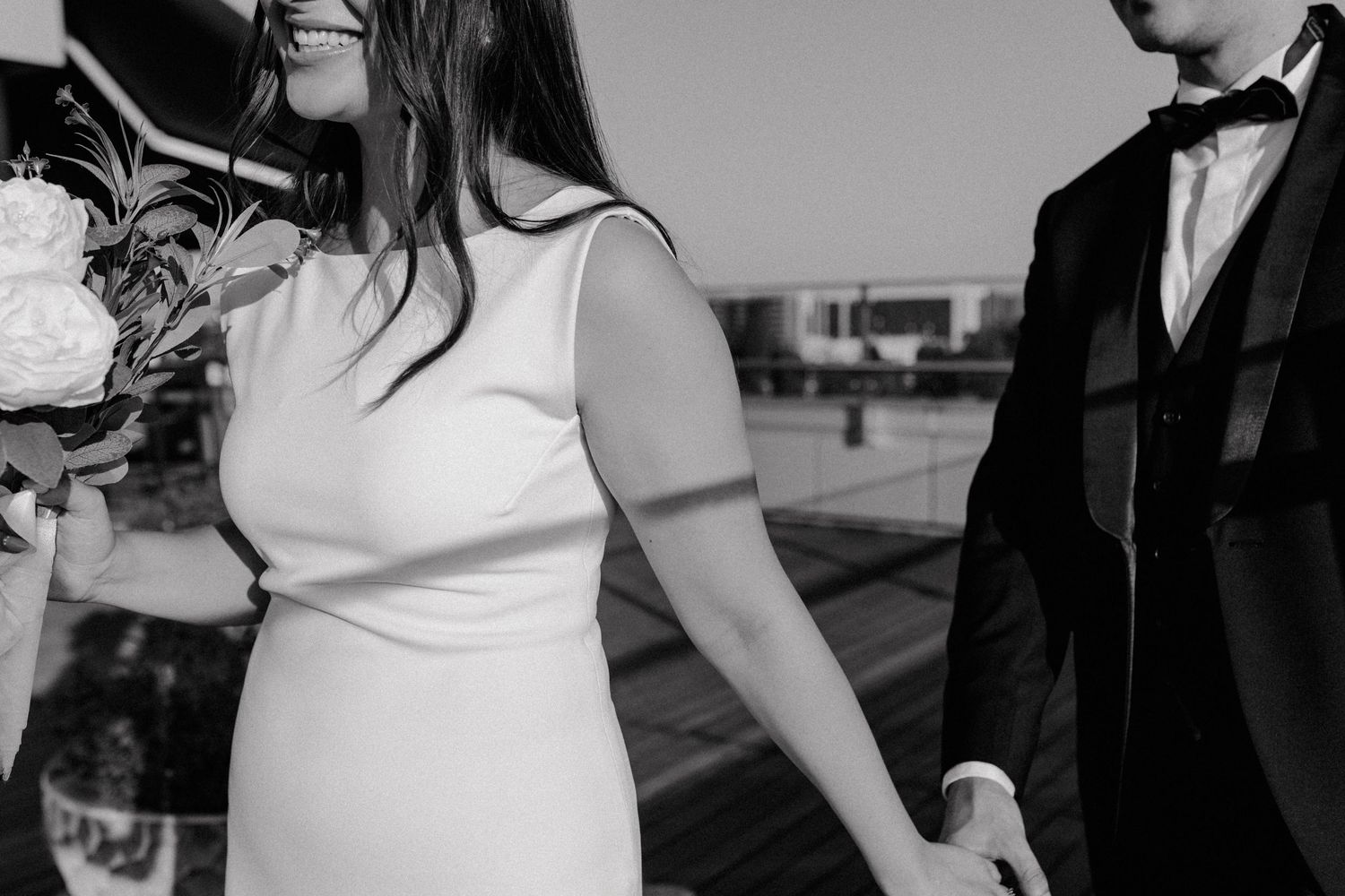 A black and white candid shot of a couple in wedding attire holding hands outdoors.