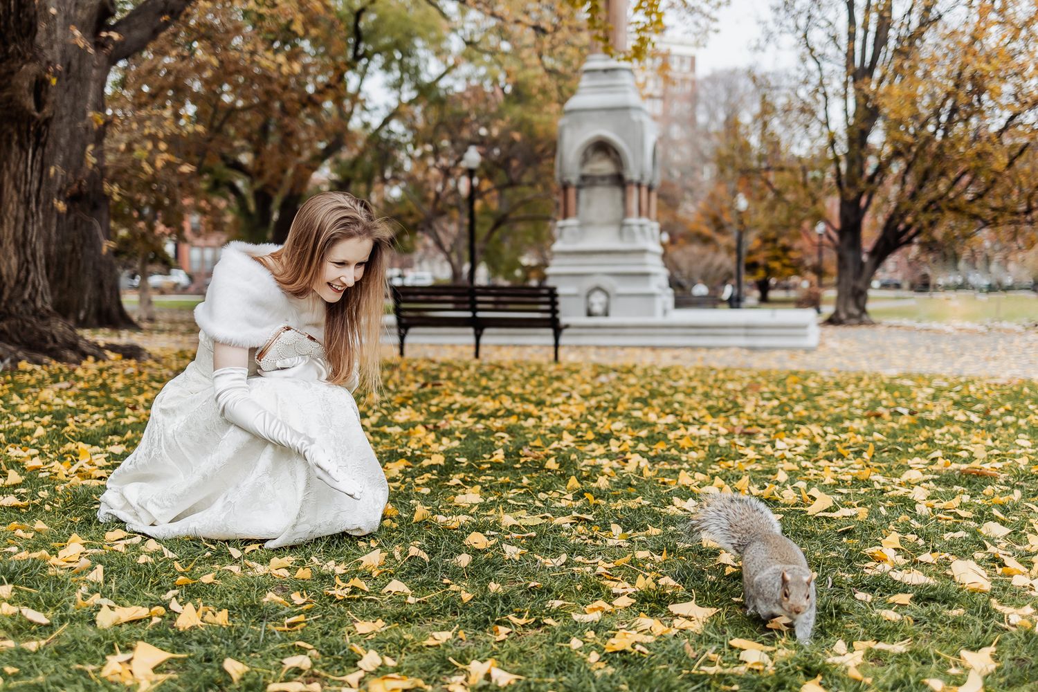 Woman in white dress crouching near a squirrel in an autumn park with yellow leaves and a monument.