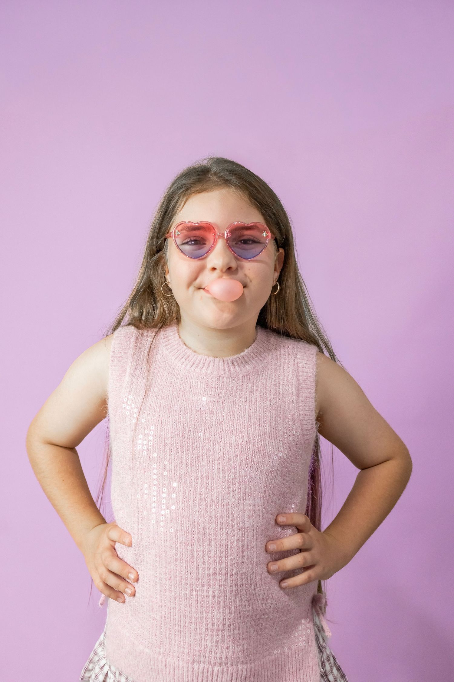 Playful young girl in pink sequin top blows bubble gum wearing heart-shaped sunglasses on purple background.