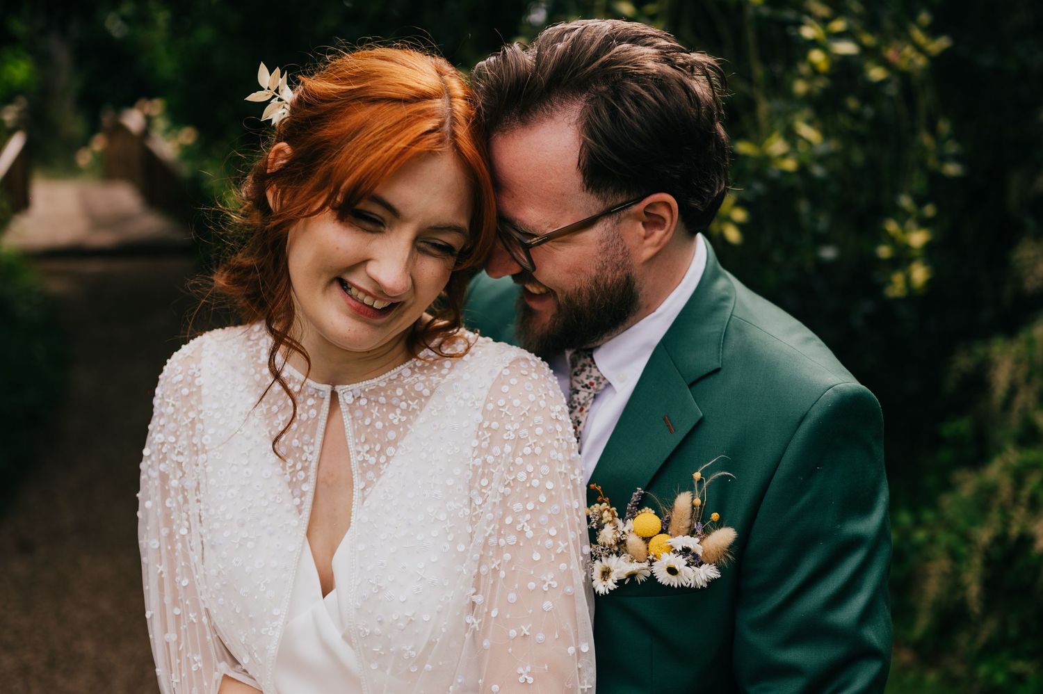 Bride and groom sit together on a bench surrounded by lush green trees in a romantic garden wedding portrait.