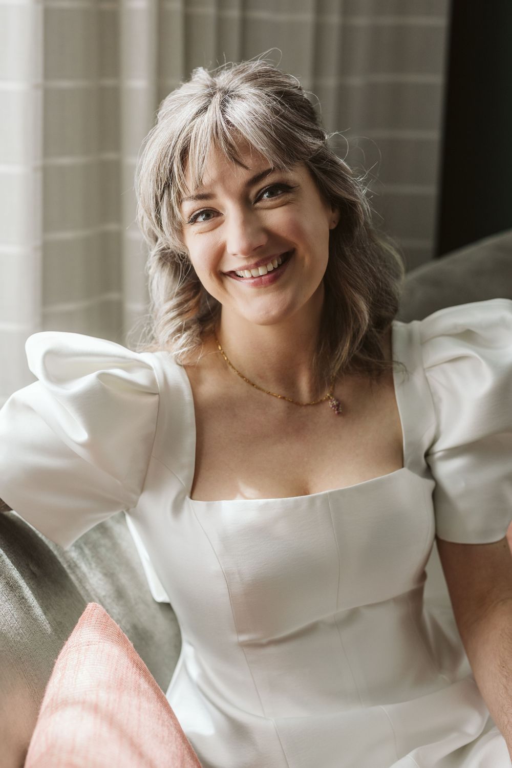 Smiling woman with silver hair wearing a white puff-sleeve dress and gold necklace, seated on a sofa.