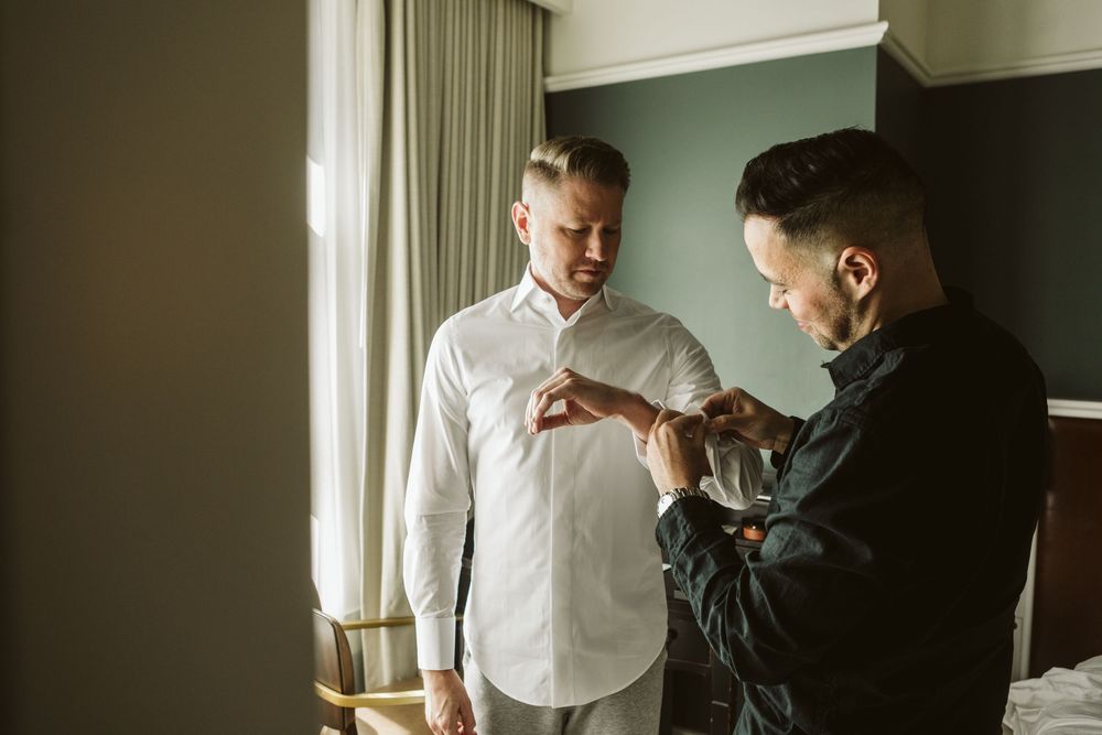 Two men in a hotel room, one in white shirt getting cufflinks fastened by another in black shirt.