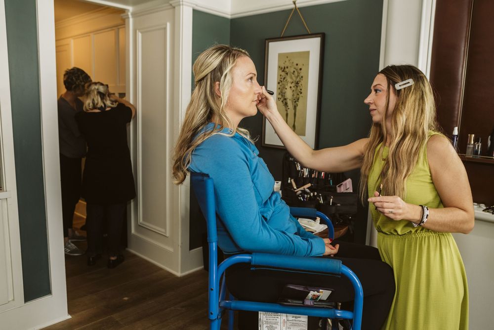 A makeup artist in a green dress applies makeup to a woman in a blue top seated in a chair indoors.