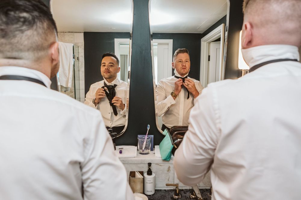 Two groomsmen adjusting ties in bathroom mirror before wedding ceremony.