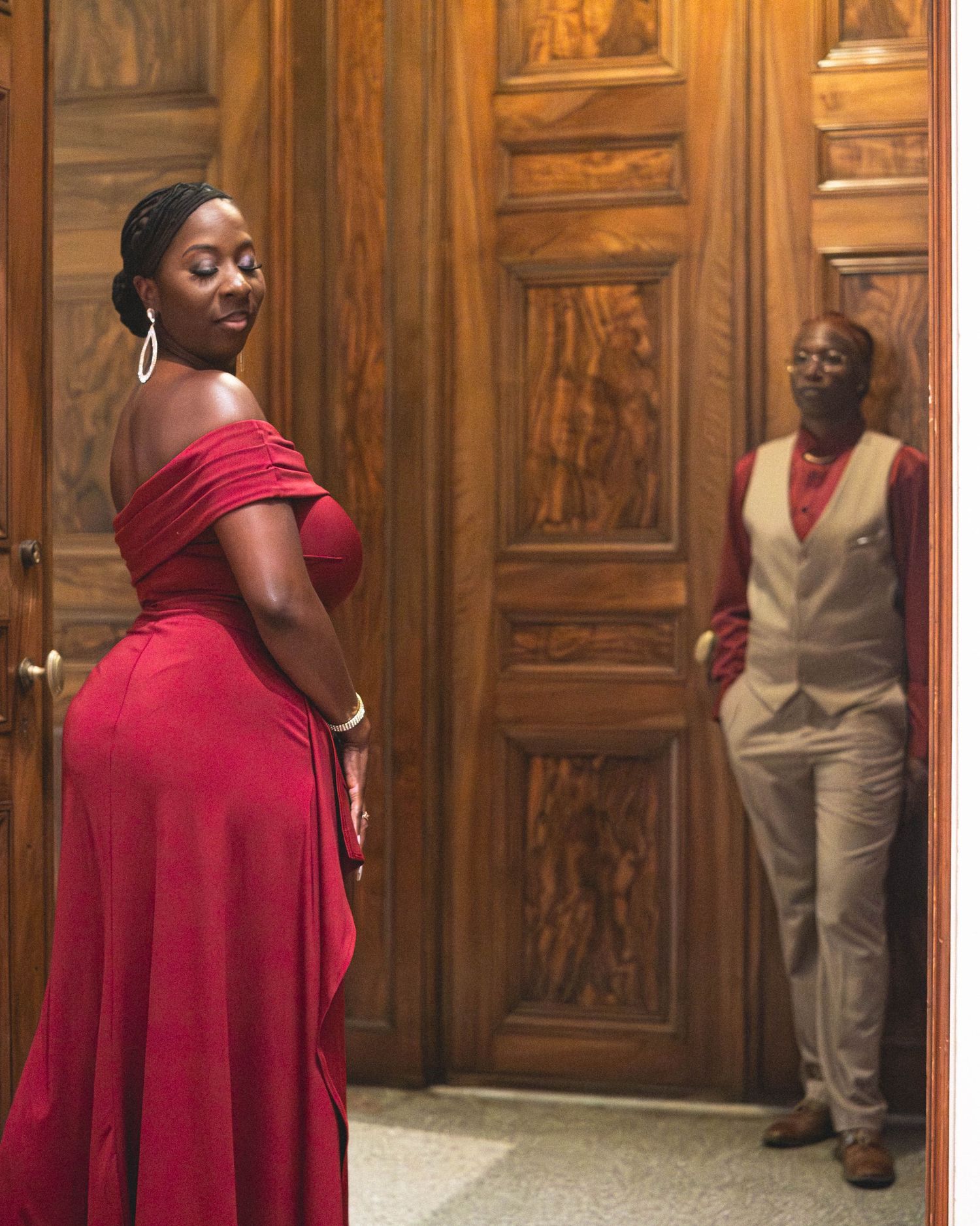 Two elegantly dressed people pose near ornate wooden doors, woman in red gown, man in tan vest.