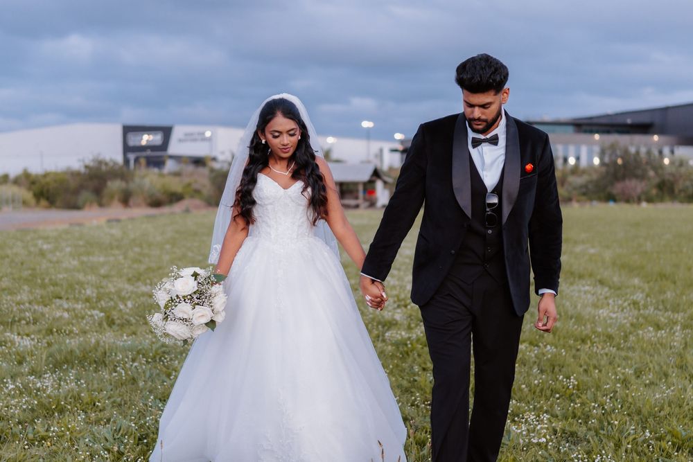 Bride in white ball gown and groom in black tuxedo hold hands walking through green field on wedding day.