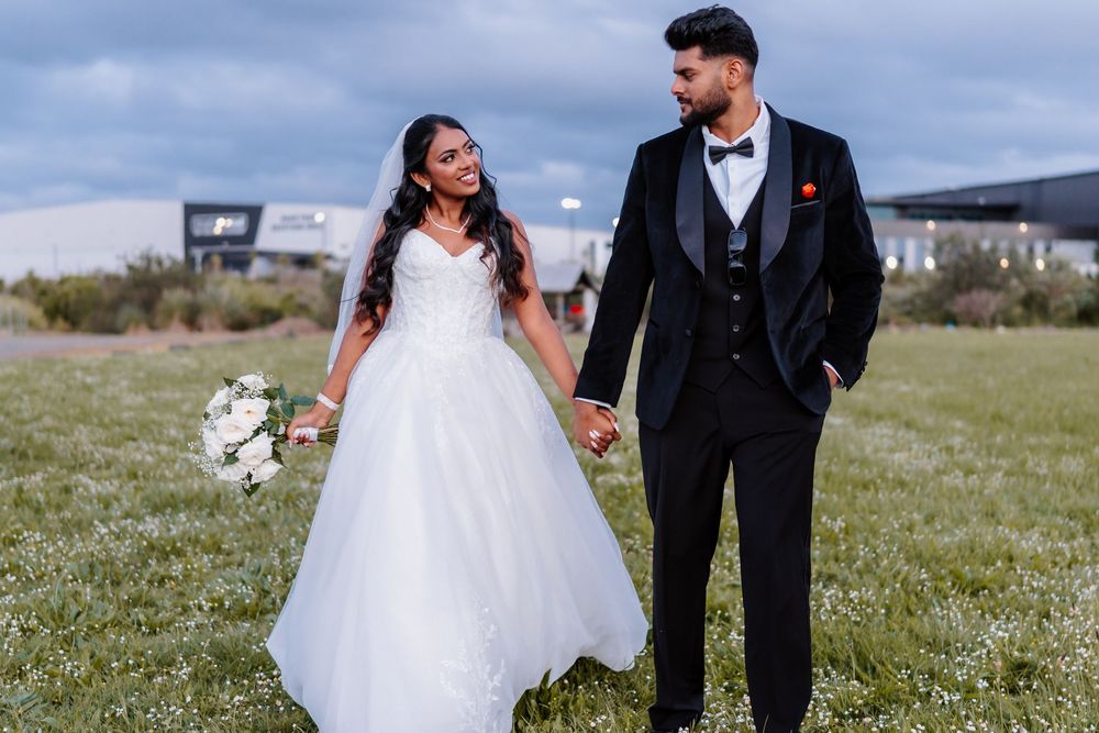 Bride and groom holding hands in a green field, she wears a white ball gown, he wears a black tuxedo.