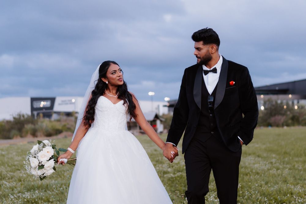 Bride and groom holding hands in a field, smiling at each other, wearing wedding attire with cloudy sky backdrop.