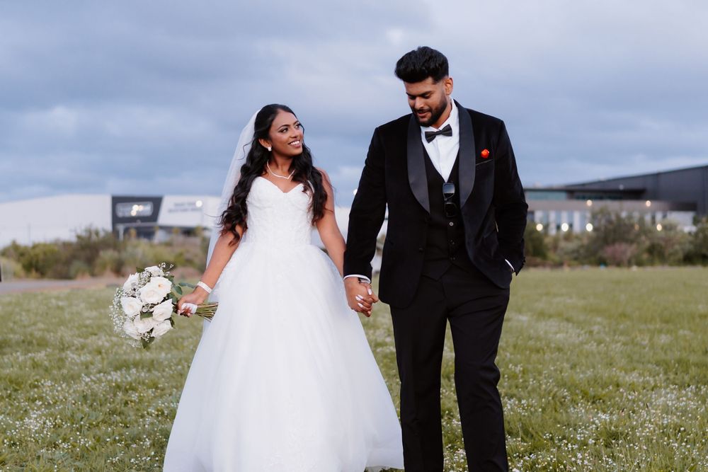 Smiling bride holding bouquet and groom in black tuxedo walking hand in hand in a field.