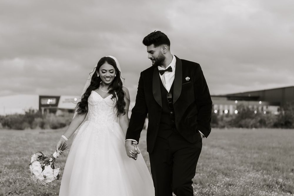 Black and white photo of a smiling bride in a ball gown and groom in a tuxedo holding hands outdoors.