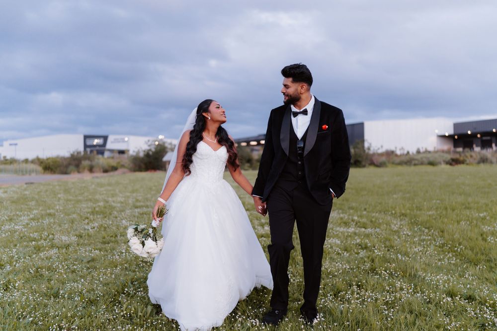Bride and groom holding hands in a green field, bride in white ballgown, groom in black tuxedo with red boutonniere.