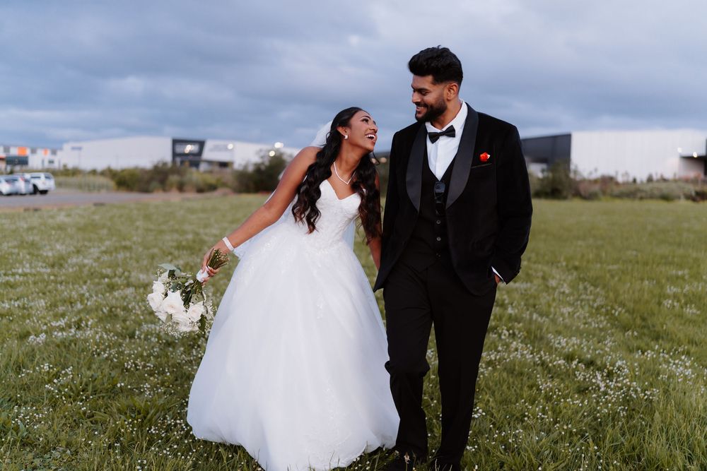 Smiling newlyweds walking through a lush green field, bride in white ballgown holding bouquet, groom in black tuxedo.