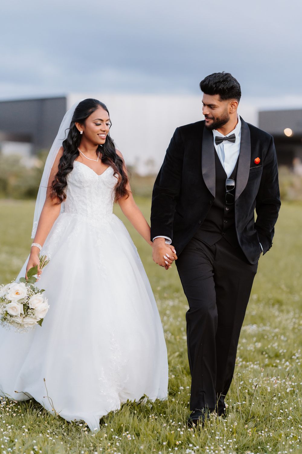 Bride in white ballgown and groom in black tuxedo holding hands walking through a flower field.