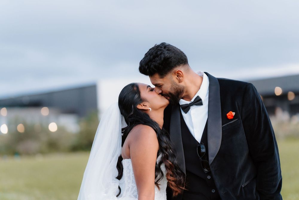 Bride and groom sharing a romantic kiss outdoors, wearing wedding attire with veil and black tuxedo with red boutonniere.
