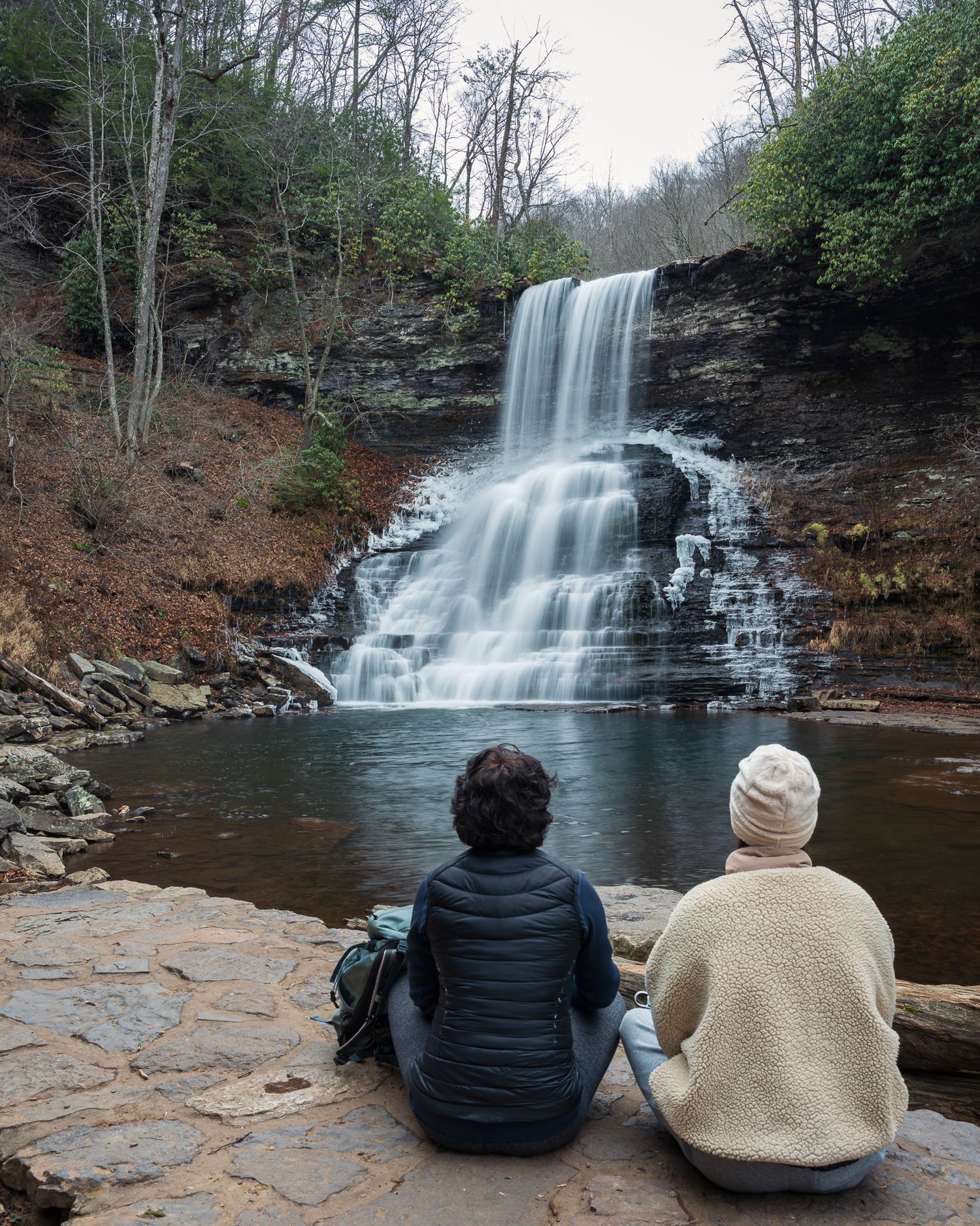 Two people sitting on rocks admiring a cascading waterfall in a lush forest canyon.