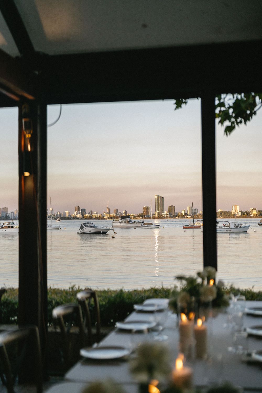Elegant waterfront dining setup with candles, white plates, and city skyline views at sunset over calm waters.