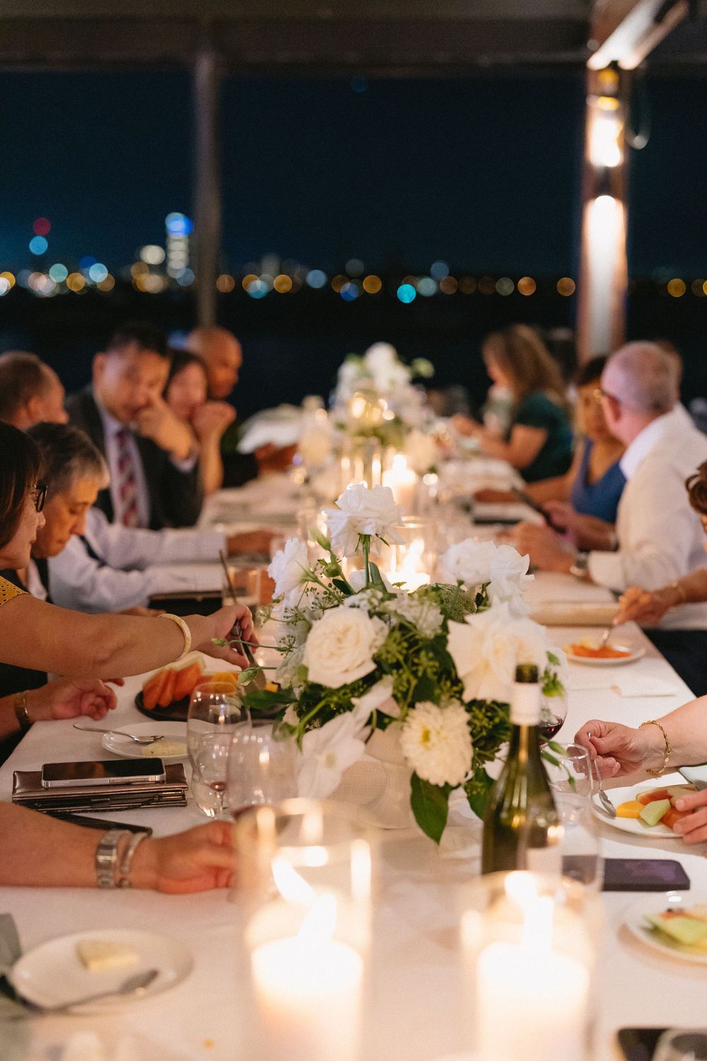 Elegant outdoor wedding reception dinner with white floral centerpieces, candles, and city lights in the background at night.