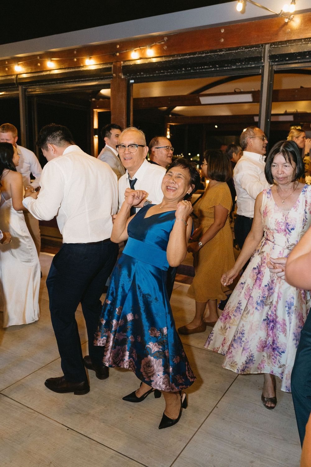 Guests dancing joyfully at a wedding reception, woman in blue floral dress smiling, elegant evening celebration.