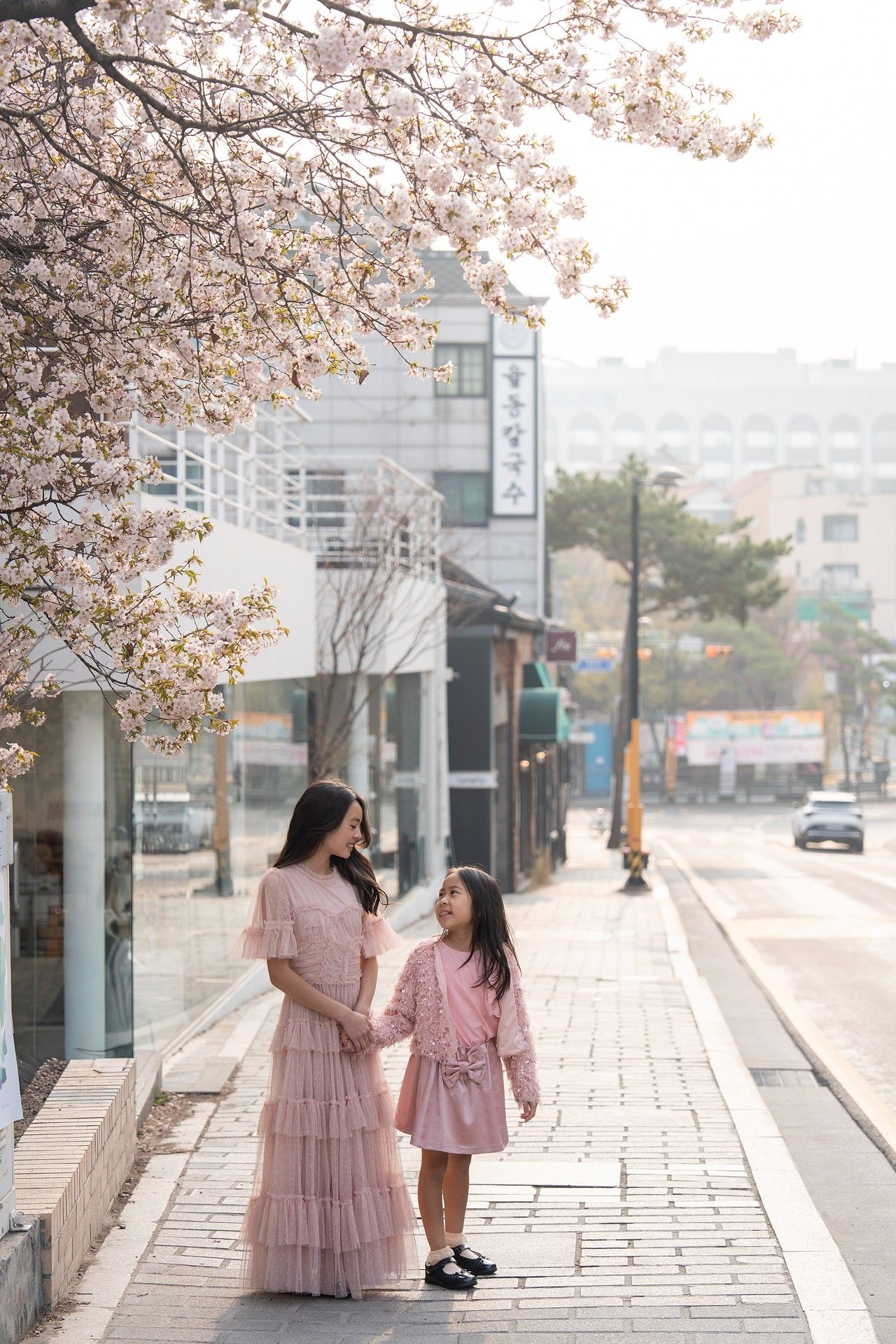 Mother and daughter in matching pink dresses stand under cherry blossom trees on a Korean street in spring.