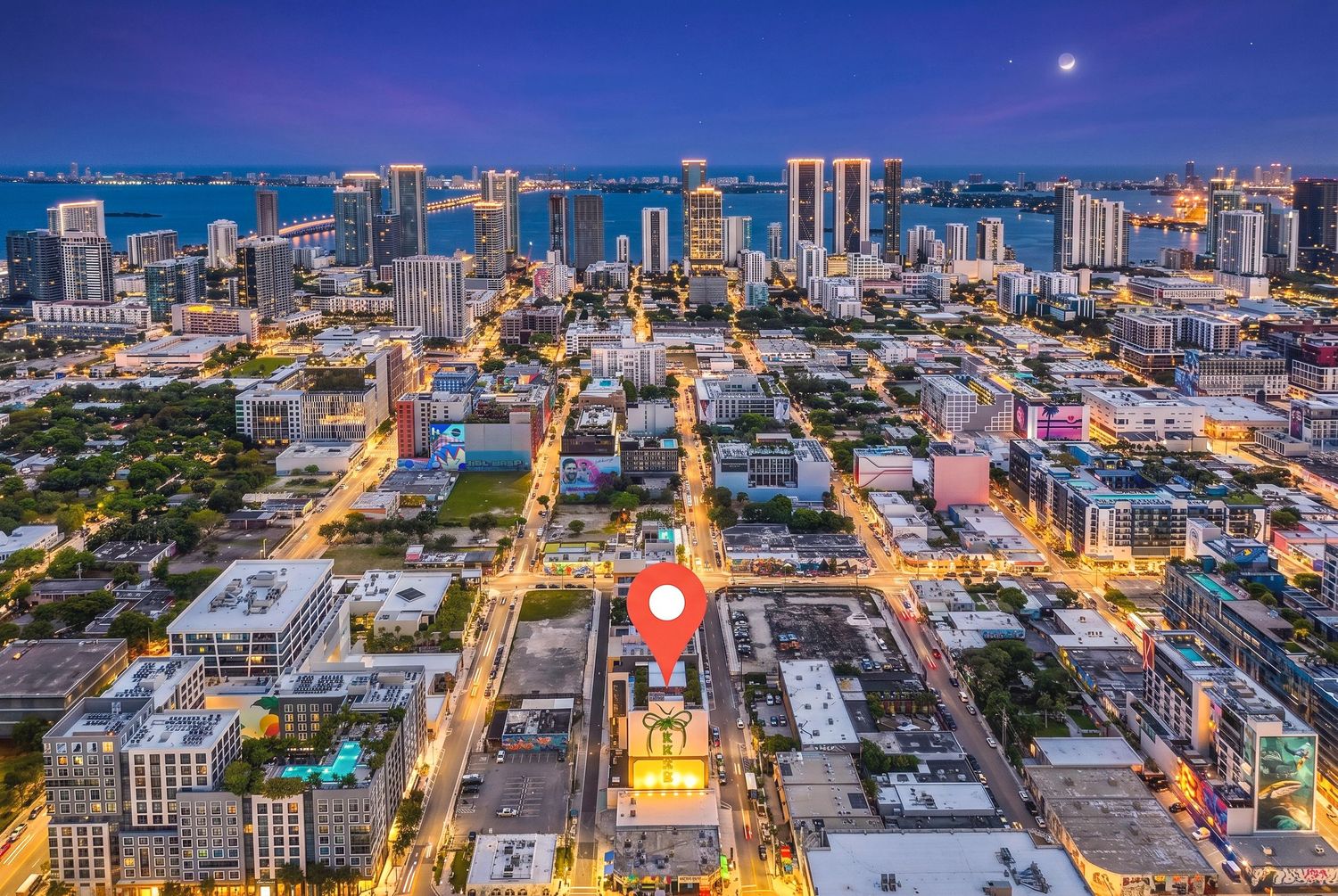Aerial twilight view of Miami cityscape with glowing streets, downtown skyline, and a location pin marker.