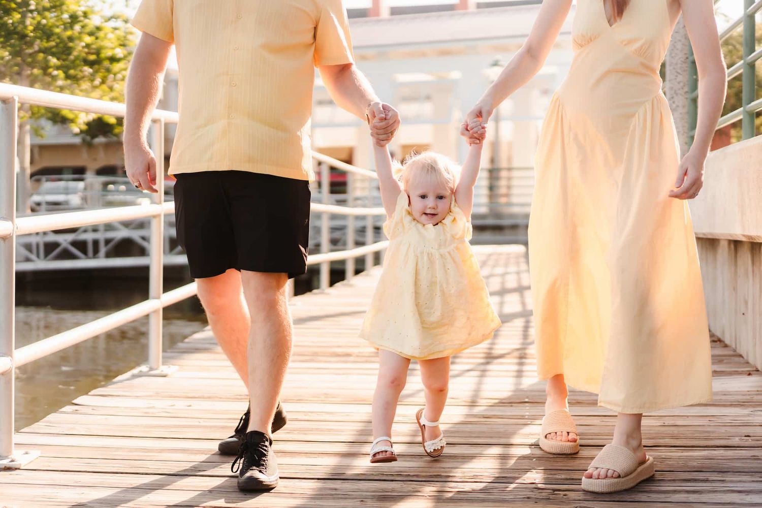 A toddler girl in a yellow dress holds hands with her parents while walking on a sunny wooden boardwalk.