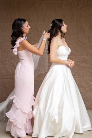 Bridesmaid in pink gown carefully adjusts the veil of bride in strapless white satin wedding dress before ceremony.