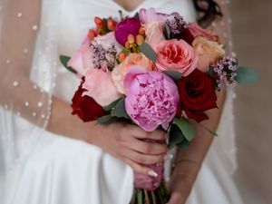 Bride holds a vibrant wedding bouquet featuring pink peonies, red and coral roses, and mixed seasonal blooms.
