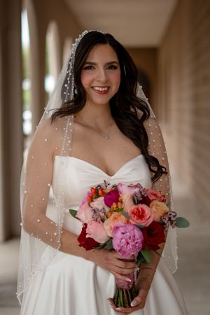 Smiling bride in white strapless gown with pearl-adorned veil holding vibrant pink and red bouquet.