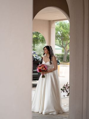 Elegant bride in white ball gown with veil holding colorful bouquet framed by arched doorway.
