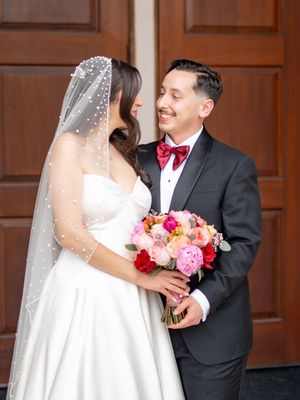 Bride and groom sharing a joyful moment, groom in black tuxedo with red bow tie at wooden doors.